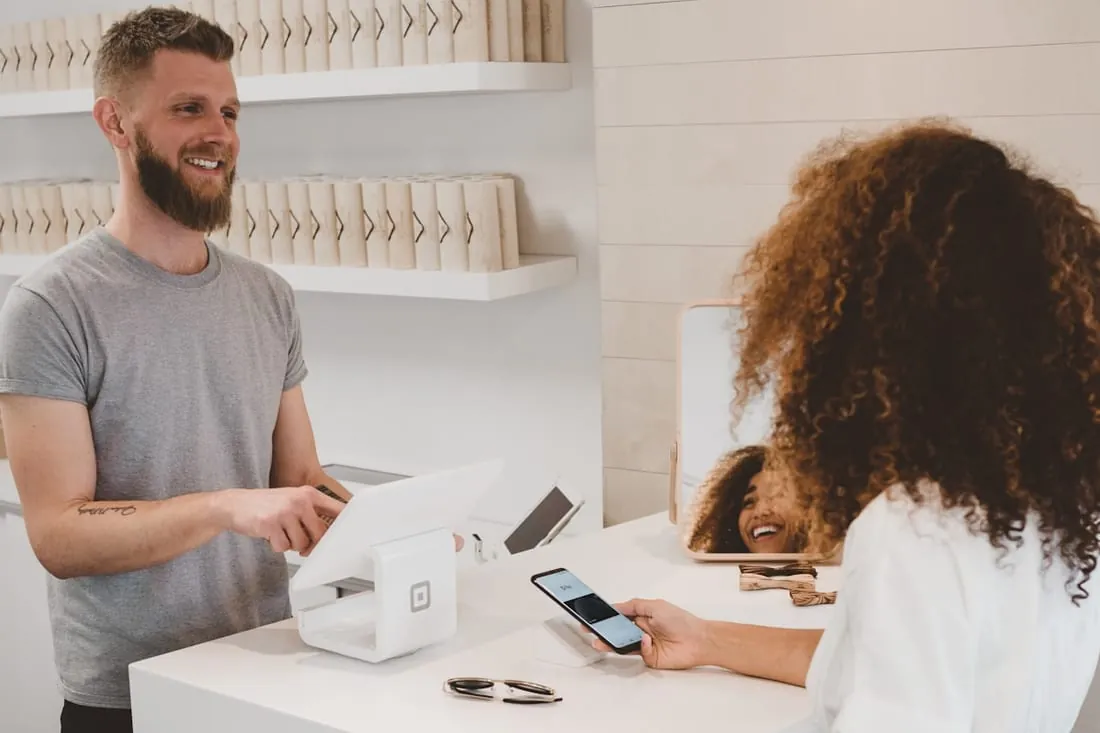 A customer pays by phone at a shop counter while the owner smiles across the till.