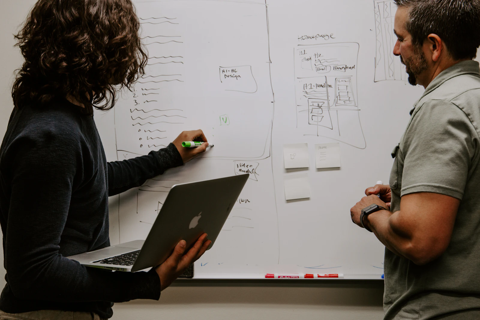 Two people planning a homepage together at a whiteboard, one holding a laptop.